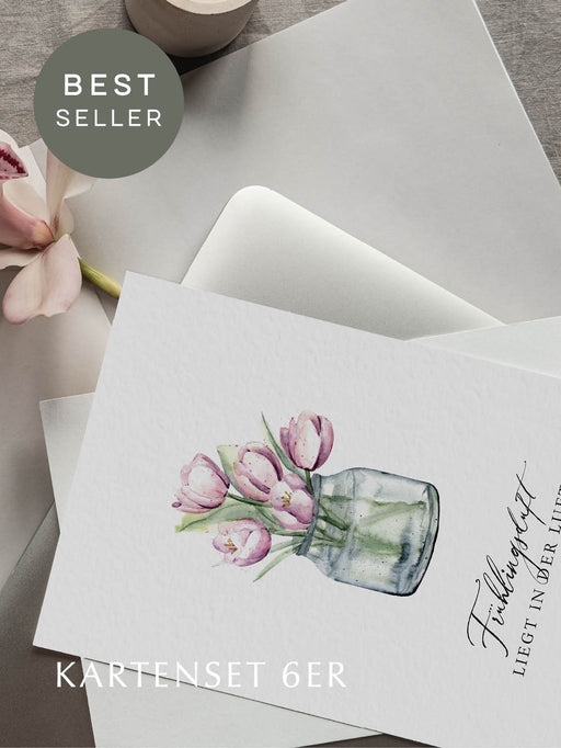 Grußkarte mit Tulpenmotiv und der Aufschrift "Frühlingsduft liegt in der Luft", liegend auf Laptop mit einer Magnolie daneben.