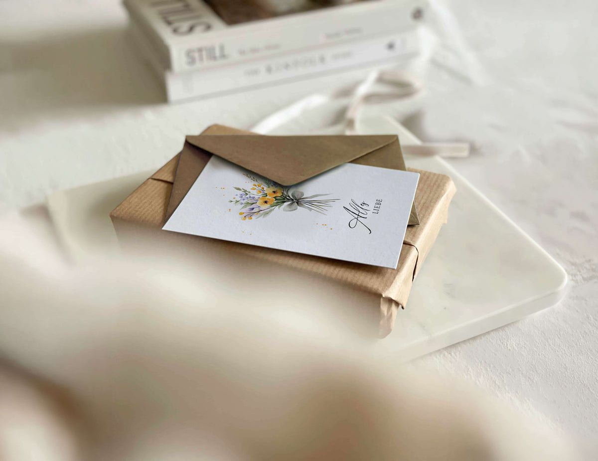 Stivolles Geschenk mit daraufliegender Grußkarte mit Blumenstrauß-Motiv und der Aufschrift Alles Liebe