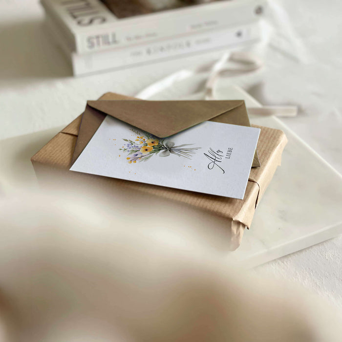 Stivolles Geschenk mit daraufliegender Grußkarte mit Blumenstrauß-Motiv und der Aufschrift Alles Liebe
