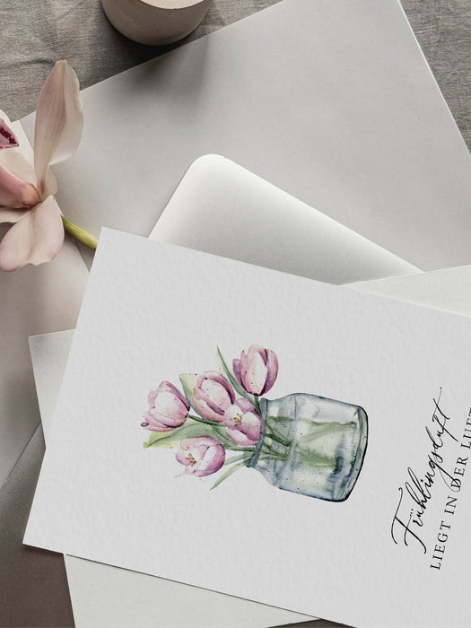 Grußkarte aus Naturkarton mit einem Tulpen-Aquarellmotiv und der Aufschrift "Frühlingsduft, liegt in der Luft" liegt auf einem Tisch mit einer Magnolie daneben. 
