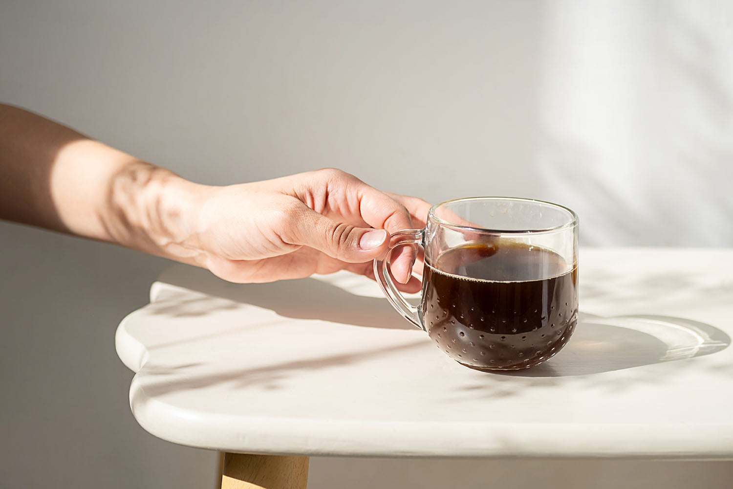 Harmonisches Bild mit einer Hand, die eine transparente Kaffeetasse auf einen weißen Tisch stellt im Sonnenschein