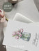 Grußkarte mit Tulpenmotiv und der Aufschrift "Frühlingsduft liegt in der Luft", liegend auf Laptop mit einer Magnolie daneben.