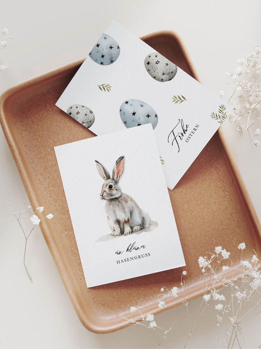 Zwei hochwertige Grußkarten zu Ostern liegend auf ein Keramiktablett mit der Aufschrift "Frohe Ostern" und "ein kleiner Hasengruß", umliegend weißem Schleierkraut
